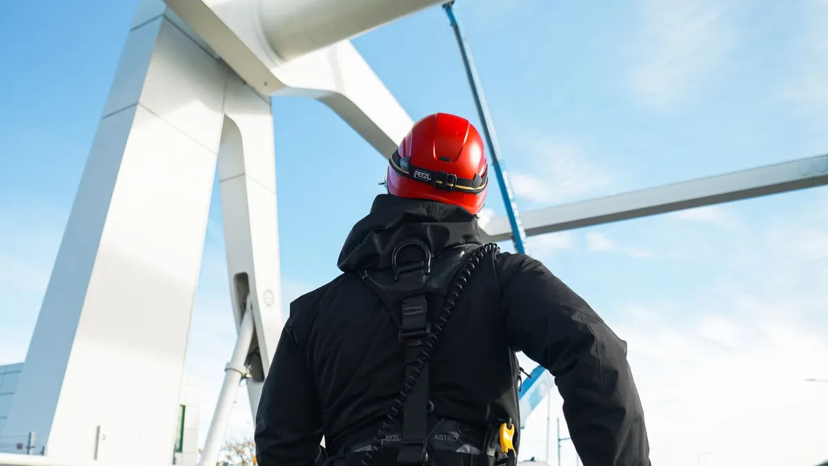 Travailleur harnaché en casque rouge, vu de dos, au pied d'une grande structure blanche